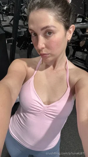 Woman in pink tank top and blue leggings taking selfie in gym.
