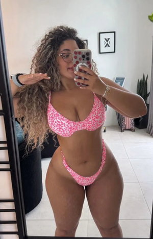 Curvy woman with curly hair in pink bikini taking mirror selfie indoors.