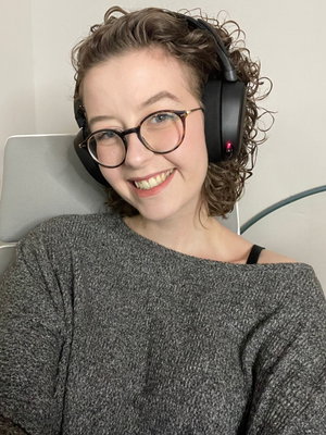 Woman with curly hair and glasses wearing headphones and a gray sweater, smiling indoors.