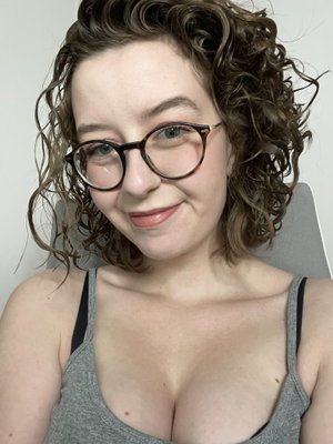 Woman with curly hair and glasses wearing a gray tank top indoors.