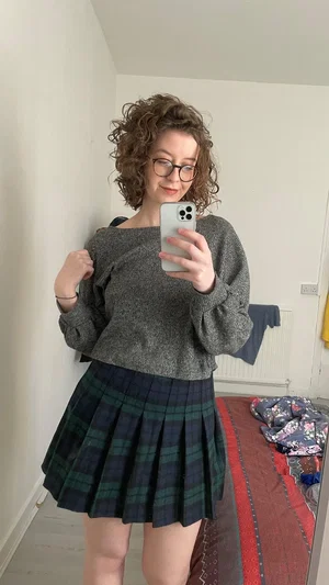 Woman with curly hair in sweater and plaid skirt takes mirror selfie in bedroom.