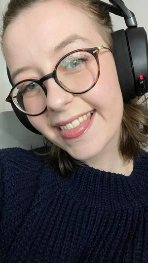 Woman with glasses and headphones, wearing a dark blue sweater, smiling in a close-up selfie.