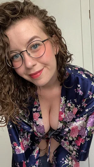 Woman with curly hair and glasses in floral robe, indoors, smiling at camera.