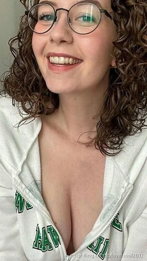Woman with curly hair and glasses, wearing a white hoodie, smiling in a close-up selfie.