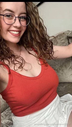 Woman with curly hair and glasses wears red tank top and white skirt, sitting on couch.