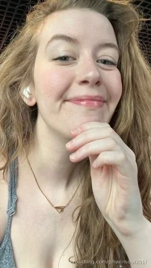 Woman with wavy hair wearing a gray tank top and gold necklace, smiling softly indoors.