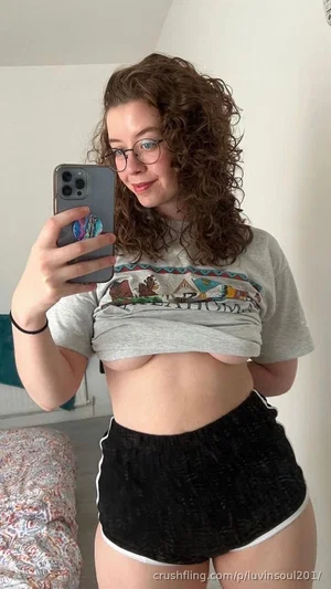 Woman with curly hair and glasses in a gray crop tee and black shorts, taking a mirror selfie in a