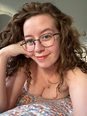 Woman with curly hair and glasses lying on floral bedding, wearing no top.