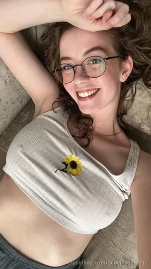 Woman with curly hair and glasses lying on couch, wearing white crop top with sunflower pin.