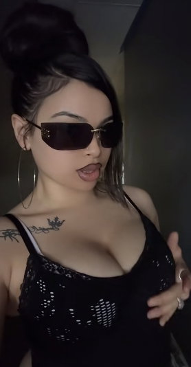 Woman with dark hair in bun, wearing black lace top and sunglasses, indoors.
