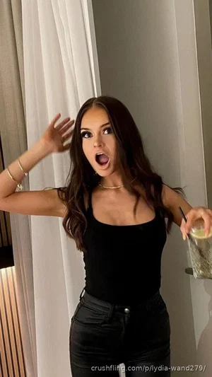 Woman with long brown hair in black tank top and black pants, holding drink, indoors.