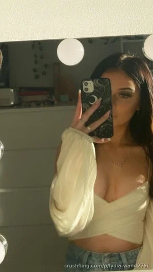 Woman in beige crop top and jeans takes mirror selfie in bedroom.
