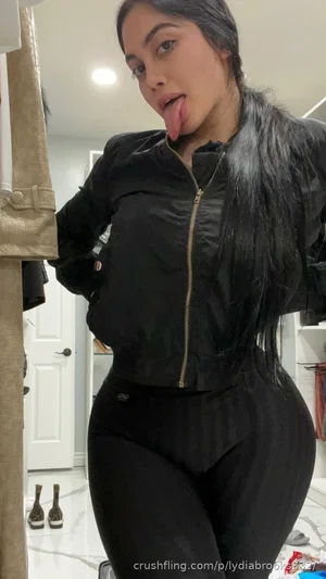 Woman with long dark hair in black jacket and leggings, posing indoors with tongue out.