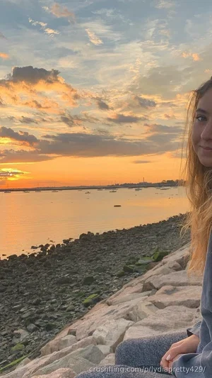 Woman with long hair sitting on rocks by water at sunset, wearing a long-sleeve top and pants.