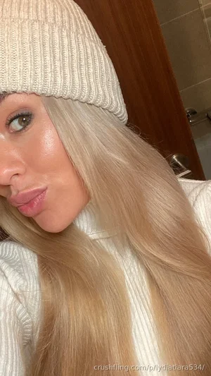 Woman with long blonde hair wearing white beanie and sweater, posing indoors.