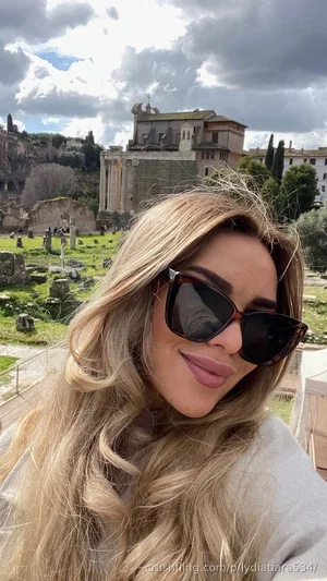 Blonde woman in sunglasses and light top, smiling in front of ancient ruins outdoors.