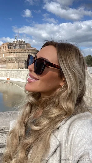 Woman with long blonde hair and sunglasses smiling outdoors near a historic building.
