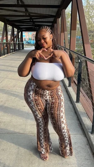 Woman in white crop top and patterned pants making heart shape with hands on bridge.