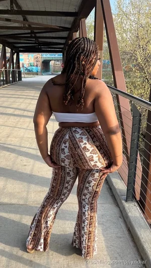 Woman with braided hair in patterned pants and white top, standing on a covered walkway outdoors.