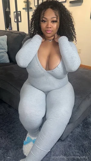 Woman in light gray ribbed bodysuit sitting on couch, curly hair, indoor setting.