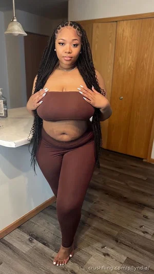 Woman with long braids in brown bodysuit standing in kitchen.