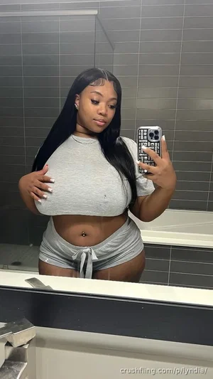 Woman with long black hair in gray crop top and shorts, taking mirror selfie in bathroom.