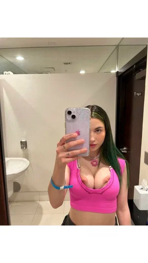 Woman with green hair in pink crop top taking mirror selfie in bathroom.