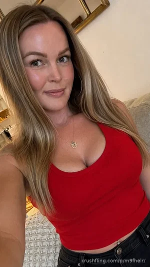 Woman with long blonde hair in red crop top and black jeans, posing indoors.