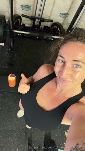 Woman in black sports bra giving thumbs up in gym, curly hair, tattoo visible.