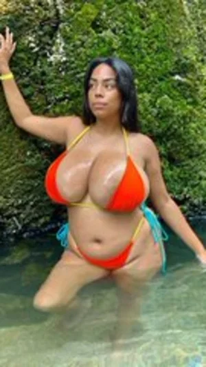 Woman in orange bikini posing in shallow water near green foliage.