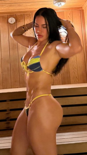 Woman in yellow and blue bikini posing in wooden sauna, showing toned physique.