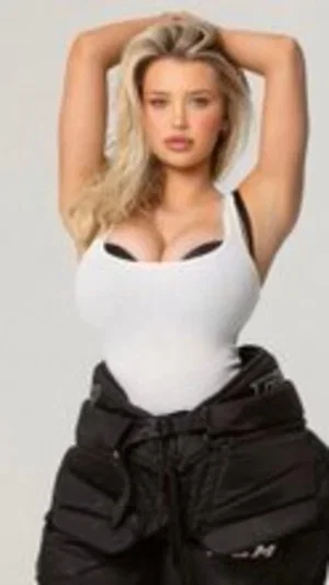Woman with blonde hair in white top and black pants, hands in hair, studio setting.
