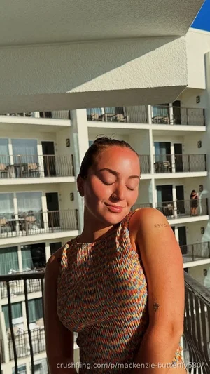 Woman in colorful knit top standing on balcony, eyes closed, smiling, with tattoos on arm.
