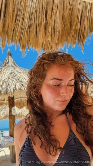 Woman with curly hair in a dark blue top under a thatched roof by the beach.