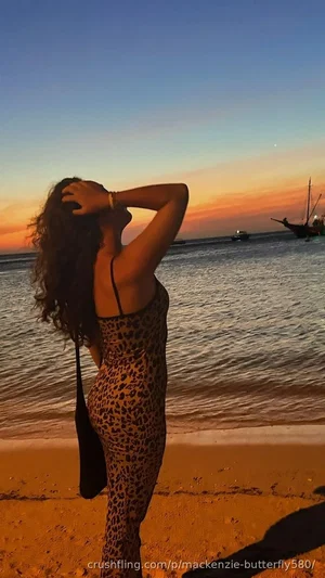 Woman in leopard print jumpsuit on beach at sunset, back to camera, hands in hair.