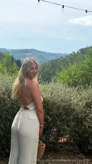 Woman with blonde hair in white backless jumpsuit, standing outdoors near greenery with hills in