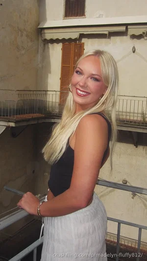 Blonde woman in black top and white skirt standing on balcony, smiling at camera.