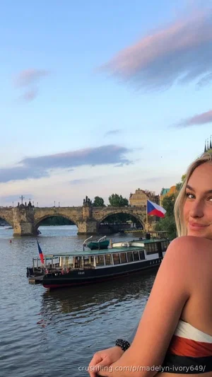 Blonde woman in strapless top posing by river with bridge and boat in background.