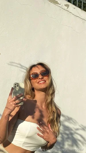 Woman with blonde hair in white crop top taking mirror selfie outdoors.