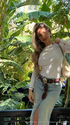 Woman in white long-sleeve top and denim skirt with slit, standing outdoors among large green