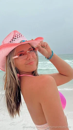 Woman in pink bikini and hat on beach, wearing sunglasses, smiling at camera.
