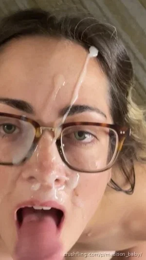 Woman with glasses, white substance on face, tongue out, indoor setting.