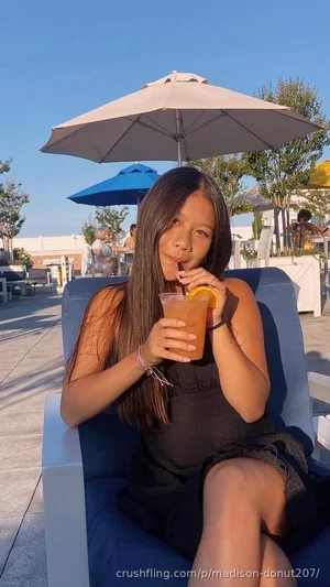 Woman with long dark hair in black dress sits on blue lounge chair outdoors, sipping drink with