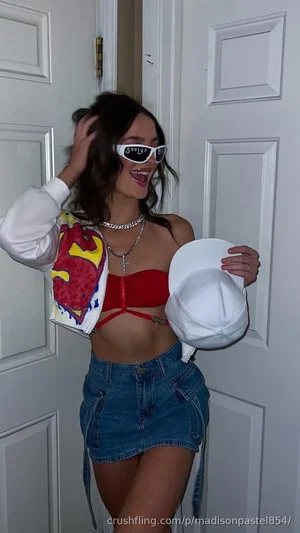 Woman in red crop top and denim skirt, holding white hat, standing by white door.