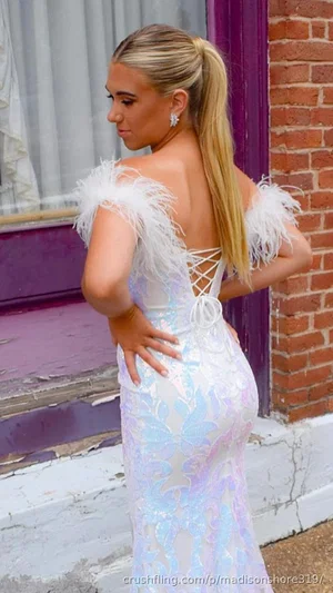 Woman in white feathered dress, blonde ponytail, standing by brick wall outdoors.