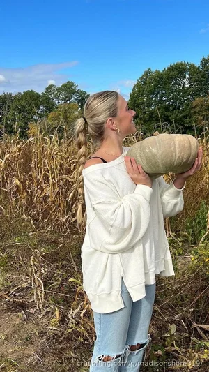 Woman with blonde braid in white off-shoulder sweater and ripped jeans holding a squash in a field.