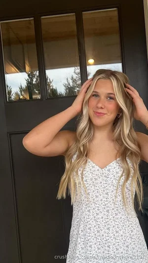 Blonde woman in floral dress standing by dark door, hands in hair, smiling.