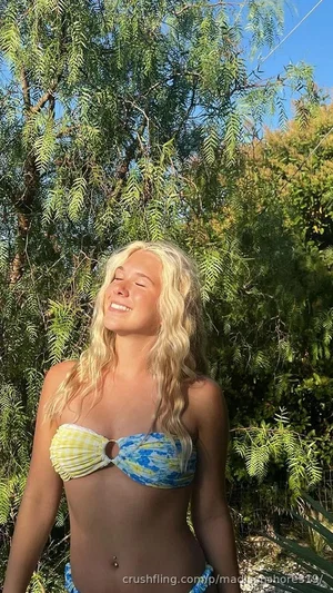 Blonde woman in colorful bikini top outdoors near green foliage, smiling with eyes closed.