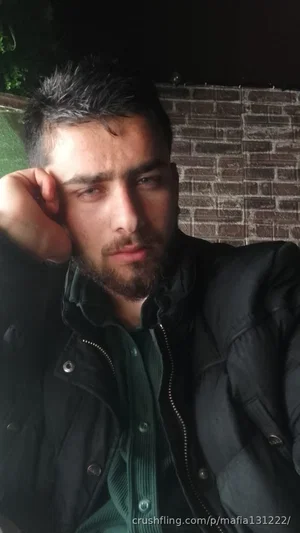 Man with short dark hair and beard wearing black jacket and green shirt, standing outdoors at night