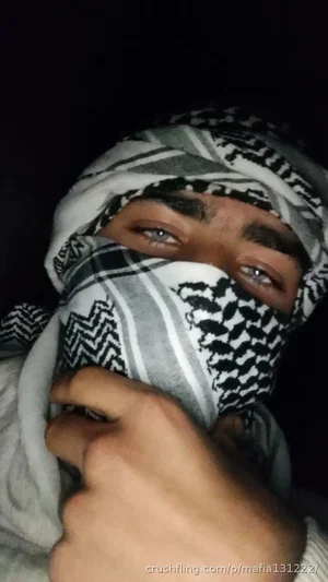 Person wearing patterned headscarf covering face, eyes visible, hand adjusting scarf.